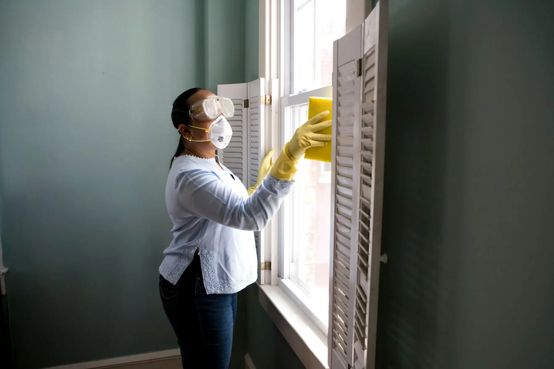 A cleaner in yellow gloves wiping a sash window in a green-painted room
