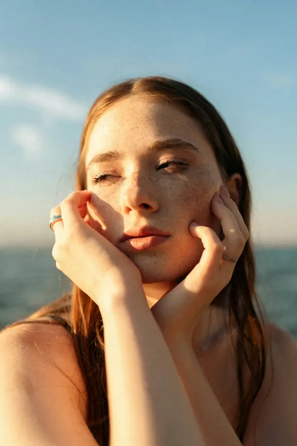 A woman with freckles in warm evening light by the sea, hands on her cheeks